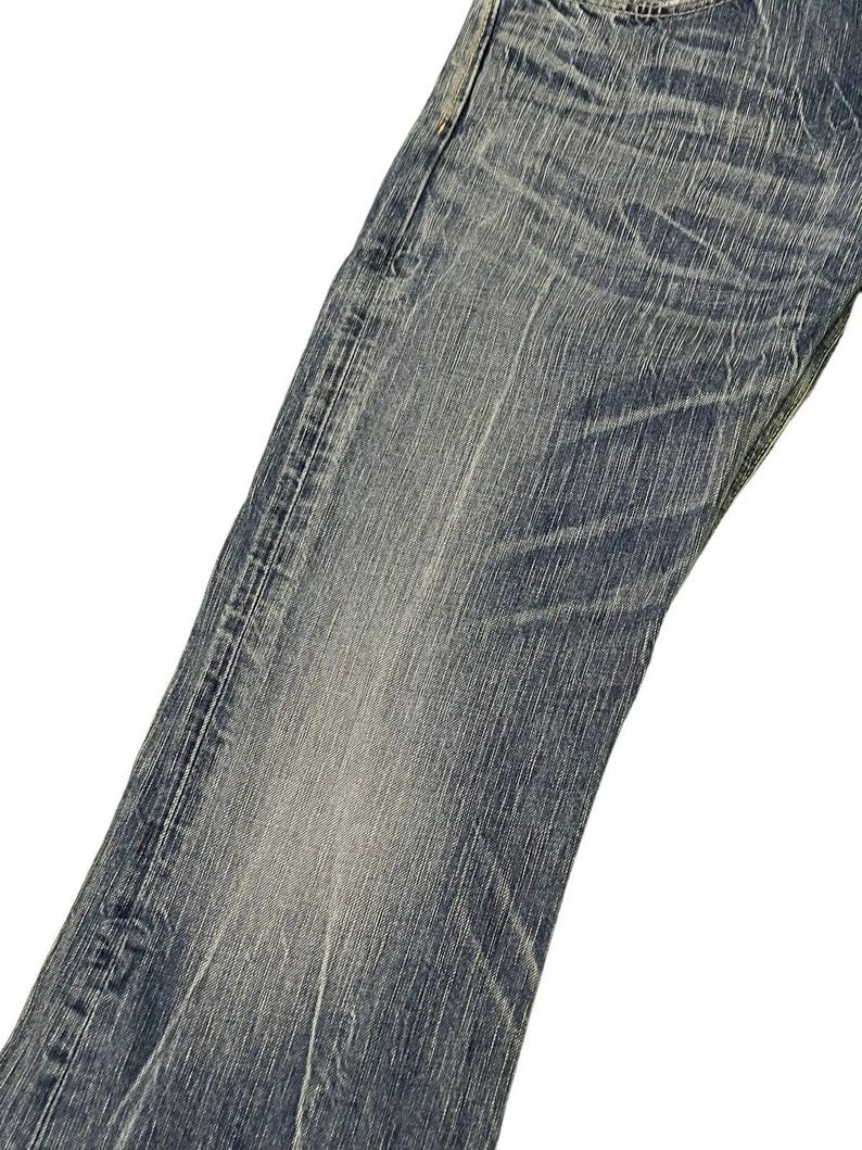 May include: Close-up of a pair of blue denim jeans. The jeans have a faded wash with visible wear and tear details. The fabric shows vertical and horizontal lines, creating a textured appearance. The jeans are angled, showing the front of the leg.
