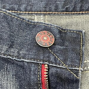 Puede incluir: Primer plano de unos vaqueros de mezclilla azul oscuro. Los vaqueros tienen una cremallera roja y un bot&oacute;n con el nombre de la marca "EDWIN" en un dise&ntilde;o circular. Las costuras son de color marr&oacute;n claro contrastante.