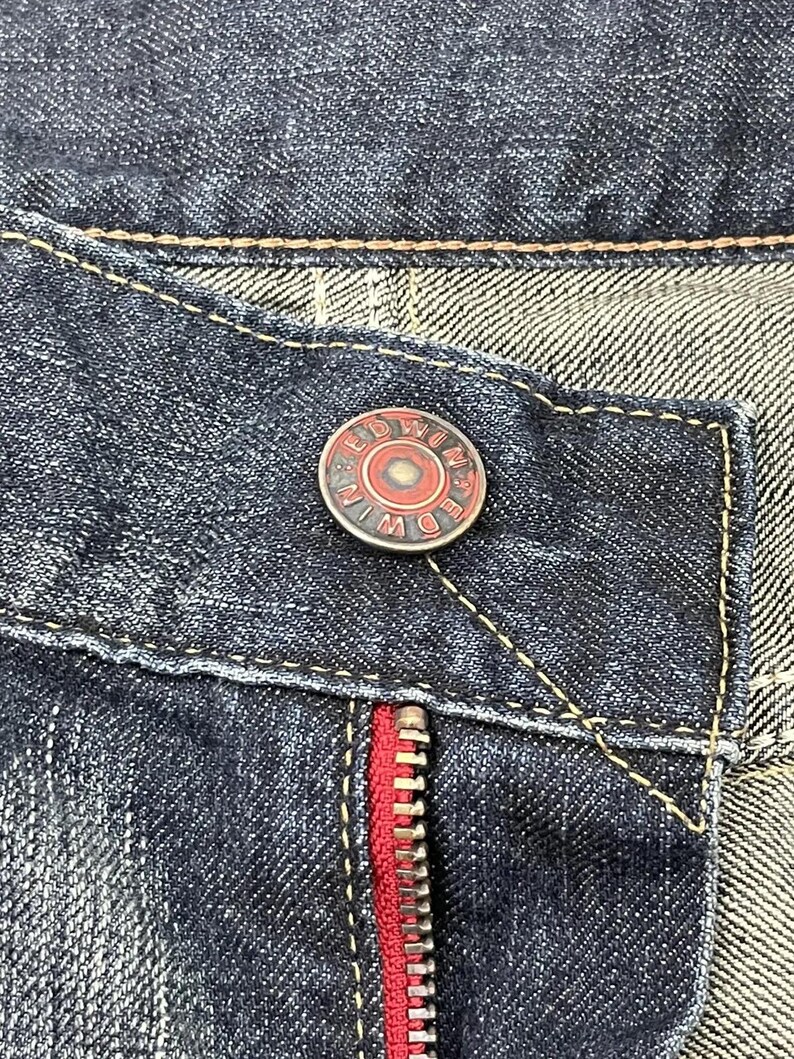 Puede incluir: Primer plano de unos vaqueros de mezclilla azul oscuro. Los vaqueros tienen una cremallera roja y un bot&oacute;n con el nombre de la marca "EDWIN" en un dise&ntilde;o circular. Las costuras son de color marr&oacute;n claro contrastante.