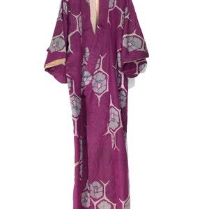 Puede incluir: Un kimono largo de color morado con un estampado floral. El kimono tiene cuello en V y mangas largas. Está colgado en una percha.