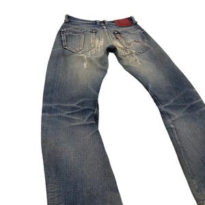 Puede incluir: Vaqueros de mezclilla azul descolorido con detalles desgastados. Los vaqueros presentan un dise&ntilde;o cl&aacute;sico de cinco bolsillos, un parche de cuero rojo y una peque&ntilde;a etiqueta roja en el bolsillo trasero. Los vaqueros tienen un ligero acampanado en la parte inferior.