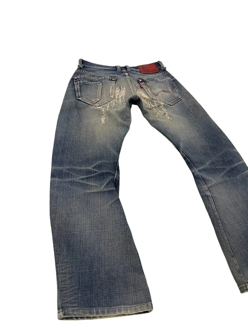 Puede incluir: Vaqueros de mezclilla azul descolorido con detalles desgastados. Los vaqueros presentan un dise&ntilde;o cl&aacute;sico de cinco bolsillos, un parche de cuero rojo y una peque&ntilde;a etiqueta roja en el bolsillo trasero. Los vaqueros tienen un ligero acampanado en la parte inferior.