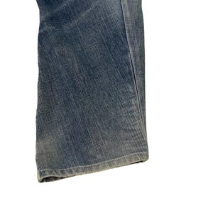 Puede incluir: Primer plano de unos vaqueros de mezclilla azules. Los vaqueros tienen un dise&ntilde;o cl&aacute;sico de cinco bolsillos y un corte de pierna recta. La mezclilla tiene un lavado descolorido, lo que le da un aspecto desgastado. Los vaqueros est&aacute;n doblados, mostrando el dobladillo y la textura de la tela.