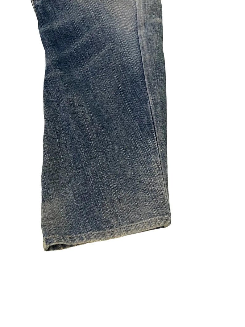 Puede incluir: Primer plano de unos vaqueros de mezclilla azules. Los vaqueros tienen un dise&ntilde;o cl&aacute;sico de cinco bolsillos y un corte de pierna recta. La mezclilla tiene un lavado descolorido, lo que le da un aspecto desgastado. Los vaqueros est&aacute;n doblados, mostrando el dobladillo y la textura de la tela.