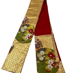 Può includere: Un lungo obi giapponese rosso e oro con un motivo floreale verde. L'obi è piegato a metà e presenta un design intrecciato.