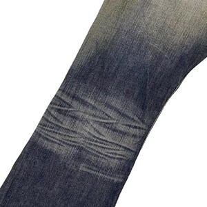 Puede incluir: Primer plano de unos vaqueros de mezclilla azules con &aacute;reas descoloridas y sutiles variaciones de textura. Los vaqueros muestran signos de desgaste, con tonos de azul m&aacute;s claros en algunas &aacute;reas, creando un aspecto vintage. El dise&ntilde;o incluye un bolsillo trasero con una costura decorativa.