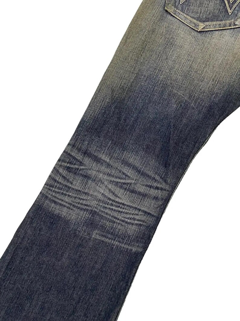 Puede incluir: Primer plano de unos vaqueros de mezclilla azules con &aacute;reas descoloridas y sutiles variaciones de textura. Los vaqueros muestran signos de desgaste, con tonos de azul m&aacute;s claros en algunas &aacute;reas, creando un aspecto vintage. El dise&ntilde;o incluye un bolsillo trasero con una costura decorativa.