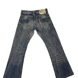 May include: Faded blue denim jeans with a bootcut leg. The jeans have a worn appearance with visible fading and distressing. A leather patch with the text "BLUE TRIP" is on the back waistband. The back pockets have a signature stitch design.