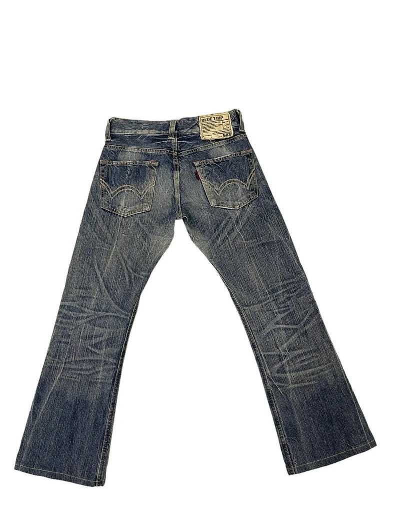 May include: Faded blue denim jeans with a bootcut leg. The jeans have a worn appearance with visible fading and distressing. A leather patch with the text "BLUE TRIP" is on the back waistband. The back pockets have a signature stitch design.