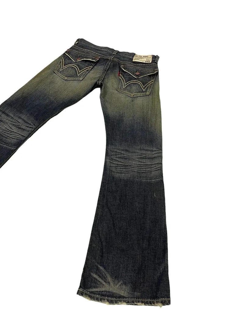 Puede incluir: Vaqueros de mezclilla azul descolorido con dise&ntilde;o de pierna acampanada. Los vaqueros presentan costuras detalladas, bolsillos traseros decorativos y una peque&ntilde;a etiqueta roja. La mezclilla tiene un aspecto desgastado.
