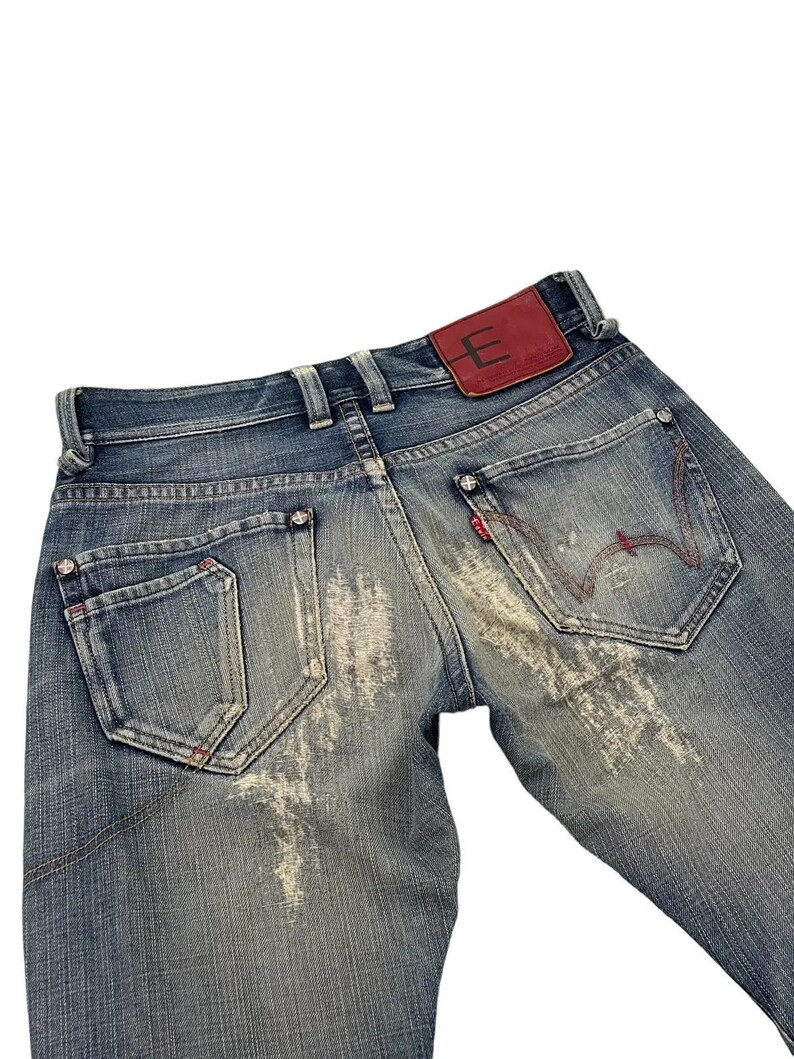 Puede incluir: Vaqueros de mezclilla azul descolorido con un parche de cuero rojo con un logotipo "E" blanco. Los vaqueros tienen detalles desgastados, costuras rojas y un dise&ntilde;o cl&aacute;sico de cinco bolsillos.