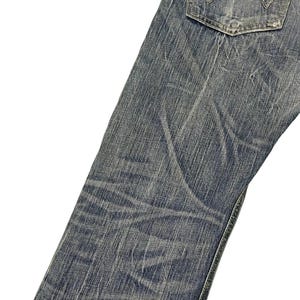 May include: Close-up of blue denim jeans with a faded wash and visible wear. The jeans feature a back pocket with a distinctive stitch design. The fabric shows creases and subtle variations in color, suggesting a worn appearance.