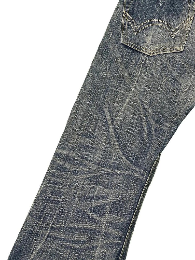 May include: Close-up of blue denim jeans with a faded wash and visible wear. The jeans feature a back pocket with a distinctive stitch design. The fabric shows creases and subtle variations in color, suggesting a worn appearance.
