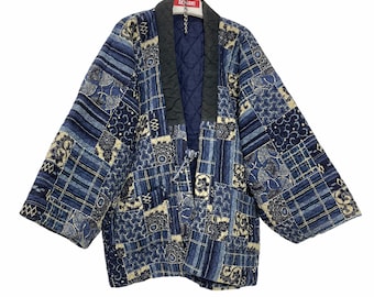 Puede incluir: Una chaqueta japonesa acolchada azul y blanca con un diseño de patchwork. La chaqueta tiene un cuello azul oscuro y está colgada de una percha.