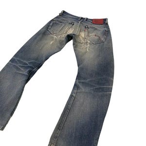 Puede incluir: Vaqueros de mezclilla azul descolorido con un dise&ntilde;o desgastado. Los vaqueros tienen un dise&ntilde;o cl&aacute;sico de cinco bolsillos, un parche de cuero rojo y una abertura de pierna acampanada. Los vaqueros est&aacute;n ligeramente desgastados y tienen un aspecto vintage.