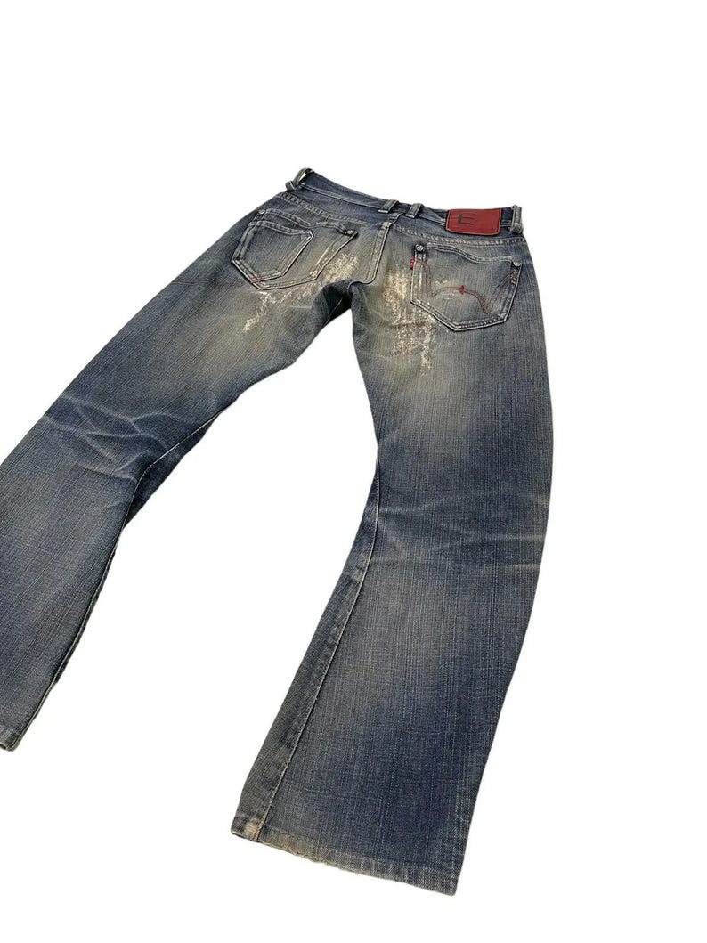 Puede incluir: Vaqueros de mezclilla azul descolorido con un dise&ntilde;o desgastado. Los vaqueros tienen un dise&ntilde;o cl&aacute;sico de cinco bolsillos, un parche de cuero rojo y una abertura de pierna acampanada. Los vaqueros est&aacute;n ligeramente desgastados y tienen un aspecto vintage.
