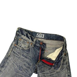 May include: Blue denim jeans with a distressed wash and a red zipper. The jeans have the number "503" printed on the waistband. The jeans are unbuttoned and unzipped, showing the interior.