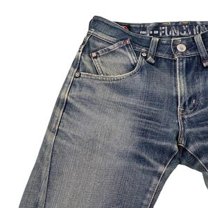 Puede incluir: Primer plano de unos vaqueros de mezclilla azules con un lavado descolorido. Los vaqueros presentan un dise&ntilde;o cl&aacute;sico de cinco bolsillos, cierre de bot&oacute;n y remaches. Las costuras son visibles y la tela tiene un aspecto texturizado. Los vaqueros est&aacute;n ligeramente desgastados.