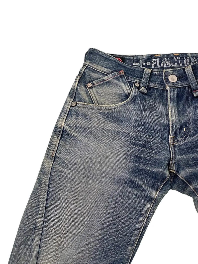 Puede incluir: Primer plano de unos vaqueros de mezclilla azules con un lavado descolorido. Los vaqueros presentan un dise&ntilde;o cl&aacute;sico de cinco bolsillos, cierre de bot&oacute;n y remaches. Las costuras son visibles y la tela tiene un aspecto texturizado. Los vaqueros est&aacute;n ligeramente desgastados.