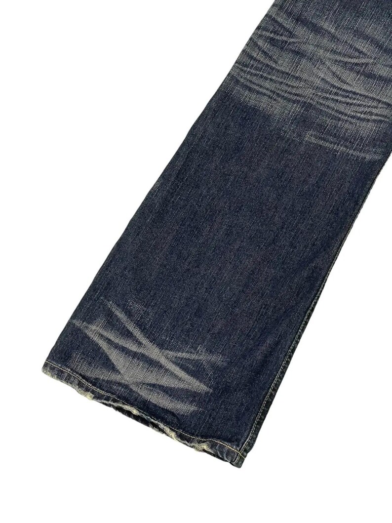 Puede incluir: Primer plano de una secci&oacute;n de vaqueros de mezclilla azul oscuro. Los vaqueros tienen un corte de pierna recta y muestran signos de desgaste con &aacute;reas descoloridas y arrugas. La mezclilla tiene una apariencia cl&aacute;sica y texturizada.
