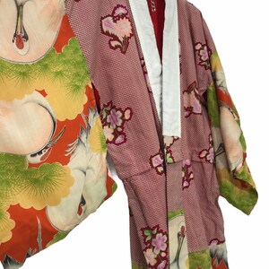 May include: A vintage kimono with a red and white crane pattern. The kimono features a red interior, white collar, and sleeves with a floral and crane design. The garment is hanging on a white hanger.