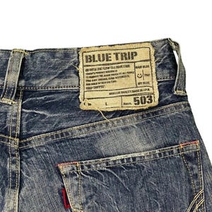 May include: Close-up of blue denim jeans with a back pocket and a label that reads "BLUE TRIP" and "503". The jeans have white stitching and a red accent on the pocket. The denim has a faded appearance.