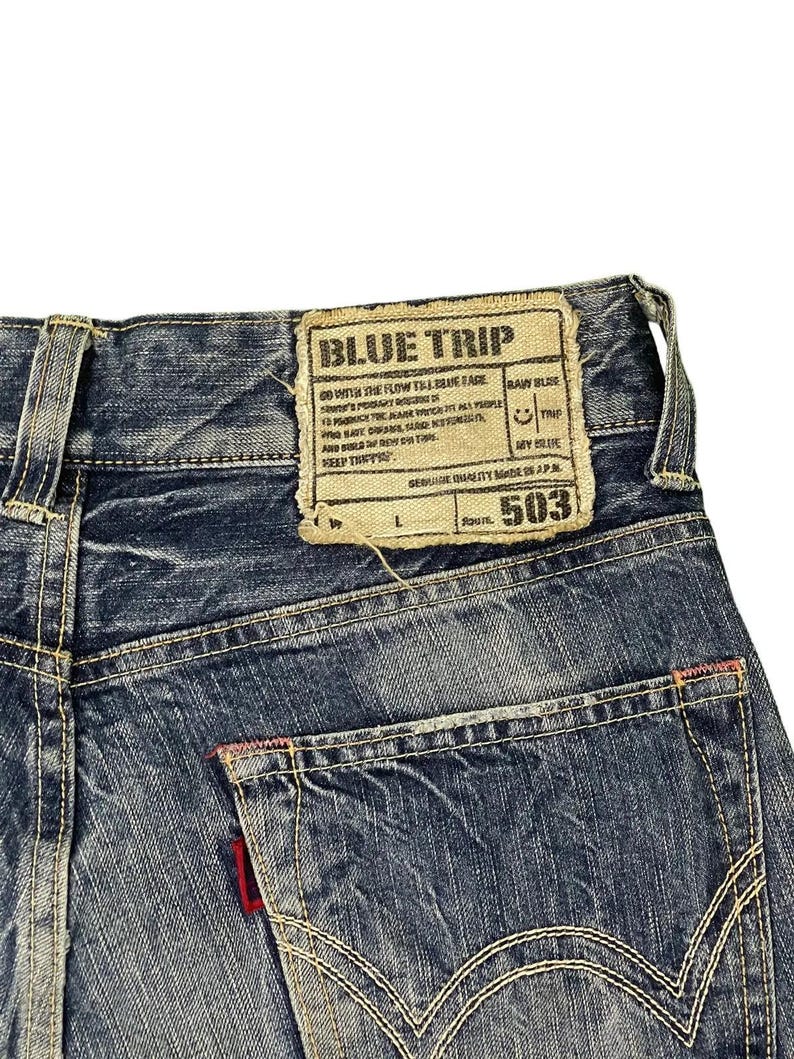 May include: Close-up of blue denim jeans with a back pocket and a label that reads "BLUE TRIP" and "503". The jeans have white stitching and a red accent on the pocket. The denim has a faded appearance.