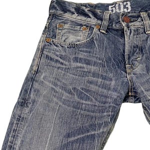 May include: Close-up of blue denim jeans with a faded wash. The jeans have a classic five-pocket design, with visible stitching and rivets. The number "503" is printed on the waistband. The denim has a worn, distressed appearance.