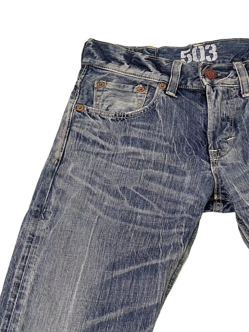May include: Close-up of blue denim jeans with a faded wash. The jeans have a classic five-pocket design, with visible stitching and rivets. The number "503" is printed on the waistband. The denim has a worn, distressed appearance.