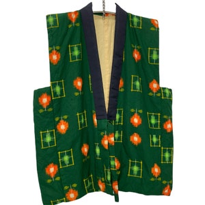 May include: A green kimono-style jacket with an orange and white floral pattern, set within yellow squares. The jacket features a navy blue collar and trim, and a cream lining. It is suspended from a metal hanger.