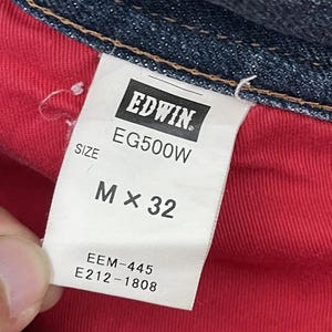 Puede incluir: Primer plano de una etiqueta blanca con la marca "EDWIN" y detalles del producto. La etiqueta muestra "EG500W" y "M x 32". Tambi&eacute;n se ven los c&oacute;digos adicionales "EEM-445" y "E212-1808". La etiqueta est&aacute; sobre un fondo de tela roja.