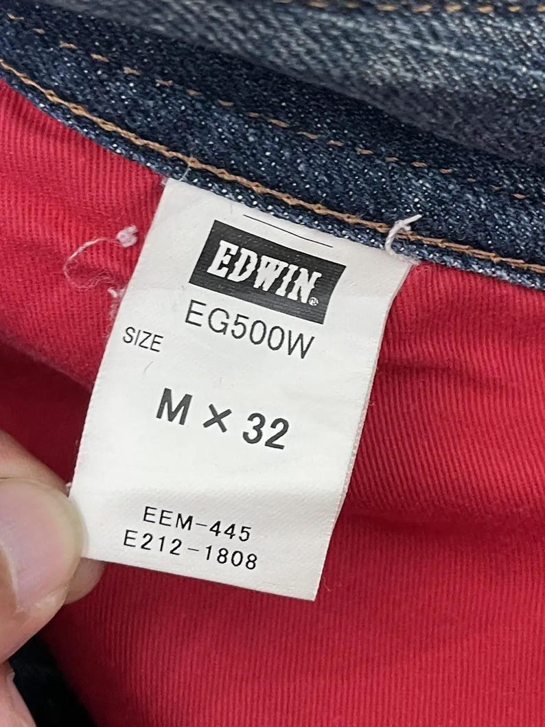 Puede incluir: Primer plano de una etiqueta blanca con la marca "EDWIN" y detalles del producto. La etiqueta muestra "EG500W" y "M x 32". Tambi&eacute;n se ven los c&oacute;digos adicionales "EEM-445" y "E212-1808". La etiqueta est&aacute; sobre un fondo de tela roja.