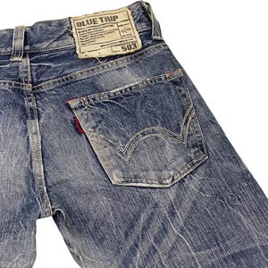 May include: Close-up of the back of blue denim jeans. The jeans feature a faded wash, a back pocket with white stitching, and a red tag. A rectangular patch with the text "BLUE TRIP" is visible.
