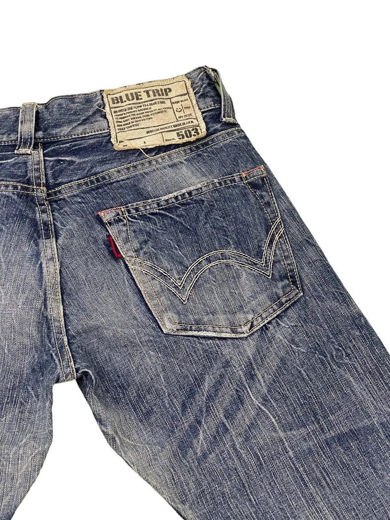 May include: Close-up of the back of blue denim jeans. The jeans feature a faded wash, a back pocket with white stitching, and a red tag. A rectangular patch with the text "BLUE TRIP" is visible.