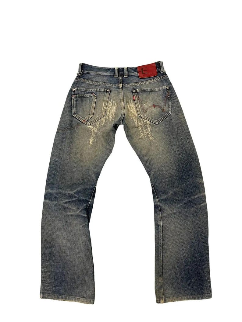 Puede incluir: Vaqueros de mezclilla azul descolorido con detalles desgastados. Incluye un parche de cuero rojo con una "E" blanca en la parte trasera y un dise&ntilde;o cosido en rojo en el bolsillo trasero. Los vaqueros tienen un dise&ntilde;o cl&aacute;sico de cinco bolsillos.