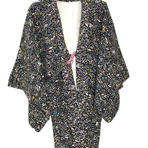 May include: A black and white patterned kimono with a pink sash. The kimono has a geometric pattern with shades of blue, yellow, and pink. The sash is tied in a bow at the front of the kimono.