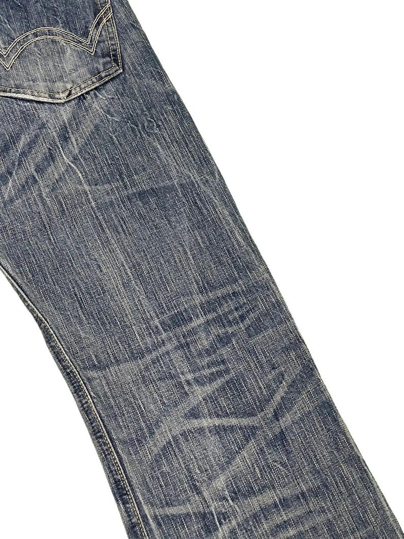 May include: Close-up of blue denim jeans with a faded, worn appearance. The jeans have a classic five-pocket design, with a visible back pocket and detailed stitching. The fabric shows signs of wear and tear, adding to the vintage aesthetic.