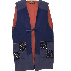 May include: A long, blue vest with a red lining and a tie closure. The vest has two large pockets with patchwork fabric insets.