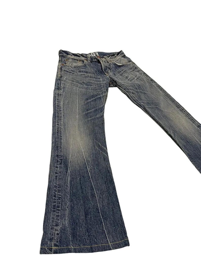 May include: Faded blue denim jeans with a flared leg design. The jeans have a worn appearance with visible fading and distressing. The stitching is visible in yellow. The jeans are laid flat against a white background.