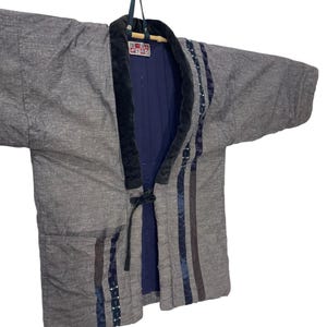 May include: Grey quilted jacket with black trim and navy blue lining. The jacket has a tie closure and decorative stripes down the front. The jacket is hanging on a hanger.