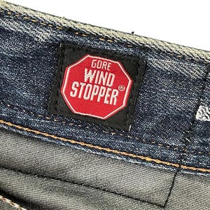 Puede incluir: Primer plano de tejido vaquero con una etiqueta negra con el texto "GORE WIND STOPPER" en letras blancas dentro de un oct&aacute;gono rojo. El tejido vaquero tiene un lavado azul y gris.