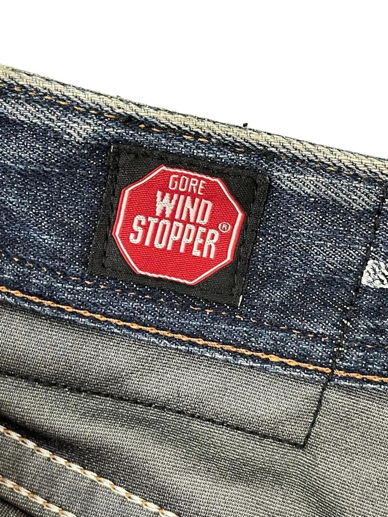 Puede incluir: Primer plano de tejido vaquero con una etiqueta negra con el texto "GORE WIND STOPPER" en letras blancas dentro de un oct&aacute;gono rojo. El tejido vaquero tiene un lavado azul y gris.