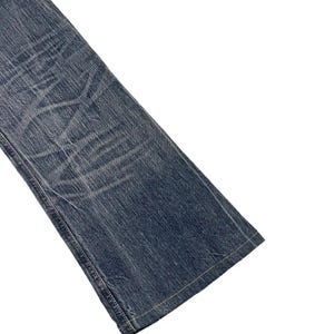 May include: Close-up of a section of blue denim jeans. The jeans have a faded, worn appearance with visible creases and subtle lighter areas. The hem and seams are visible, with a light-colored thread.