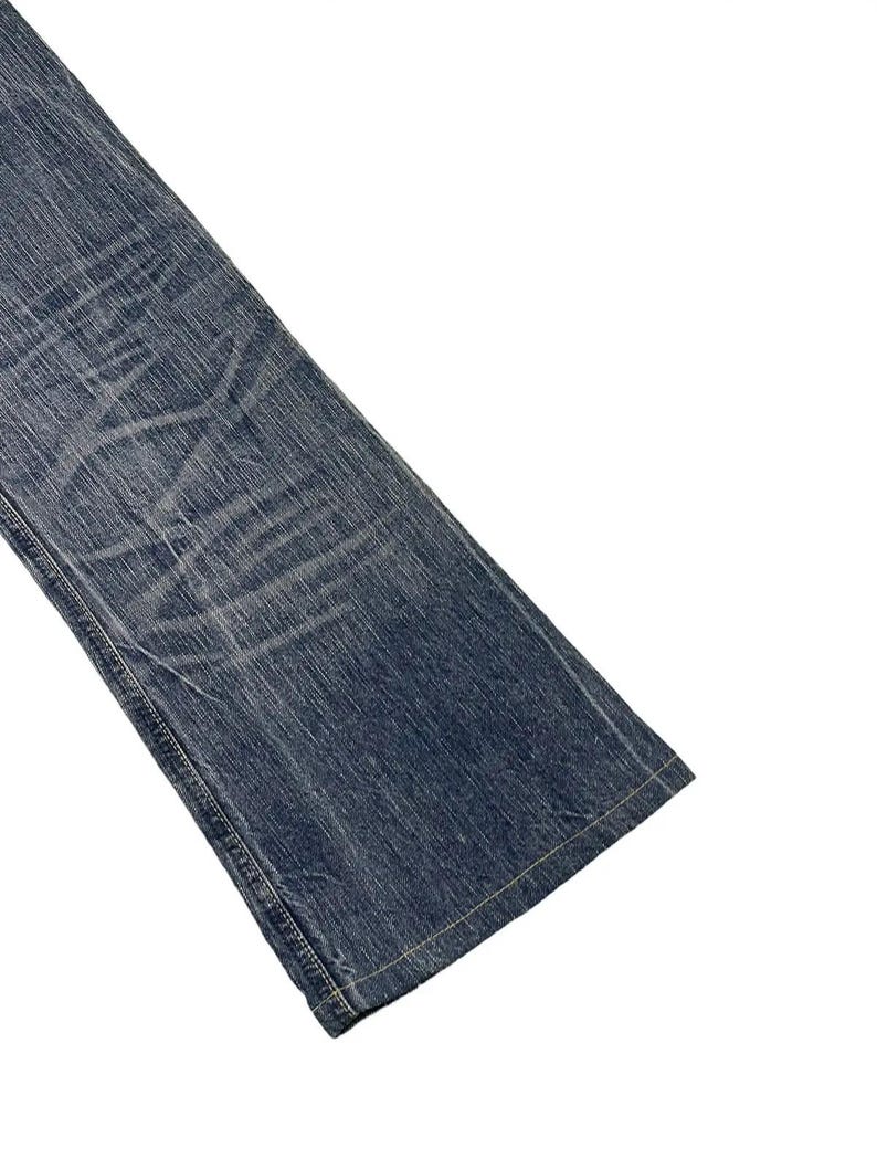 May include: Close-up of a section of blue denim jeans. The jeans have a faded, worn appearance with visible creases and subtle lighter areas. The hem and seams are visible, with a light-colored thread.