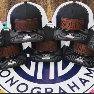May include: A collection of nine black and white trucker hats with brown leather patches featuring different names. The hats are arranged in a circle on a colorful surface with a blue and white logo in the center.