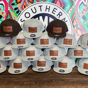 May include: A collection of gray and brown baseball caps with personalized leather patches. The caps are stacked in rows and feature various names, including Austin, Matthew, Ashton, Cason, Paxton, Braly, Jett, Rylan, Tye, Tru, Connor, Julian, Grayson, Abram, Hagen, Easton, Landon, Gunner, Koen.