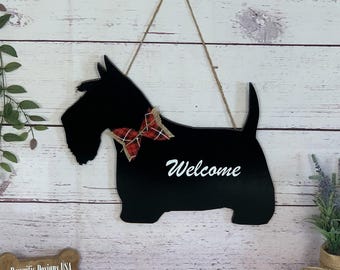 May include: Black wooden dog-shaped welcome sign with a red and black plaid bow tie. The word "Welcome" is written in white script. The sign hangs from a jute rope against a white wood background.