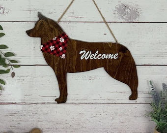 May include: Wooden dog-shaped welcome sign with a dark brown finish. The dog wears a red and black plaid bandana. The word "Welcome" is written in white script. A jute rope is used for hanging.