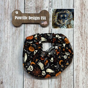 May include: A black Halloween-themed pet cone with a pattern of ghosts, pumpkins, bats, and spiderwebs. The cone is ruffled and has a matching pattern. A wooden sign with the text "Pawriffic Designs USA" is also visible.