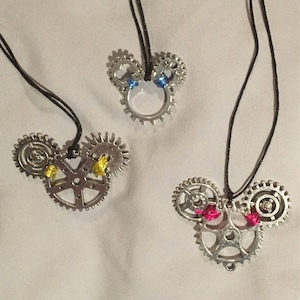 May include: Three silver necklaces with gear-shaped pendants. The pendants are shaped like Mickey Mouse heads. The first pendant has blue accents, the second has yellow accents, and the third has pink accents.